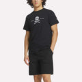 A man wears a Reebok Men's Crushers Tee in black, featuring a skull and crossed golf clubs graphic, paired with black shorts, standing with one hand in his pocket against a plain background.