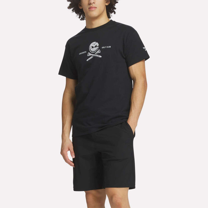 A man wears a Reebok Men's Crushers Tee in black, featuring a skull and crossed golf clubs graphic, paired with black shorts, standing with one hand in his pocket against a plain background.
