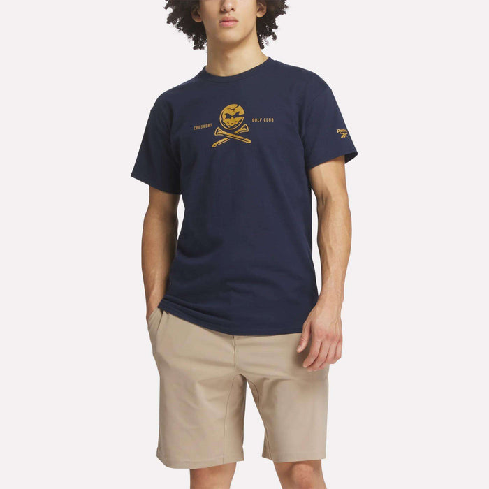 A man wears the Reebok Men's Crushers Tee in navy blue, featuring a yellow skull and crossed golf clubs graphic, paired with light beige shorts, standing against a plain white background.