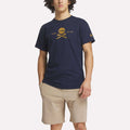A man wears the Reebok Men's Crushers Tee in navy blue, featuring a yellow skull and crossed golf clubs graphic, paired with light beige shorts, standing against a plain white background.