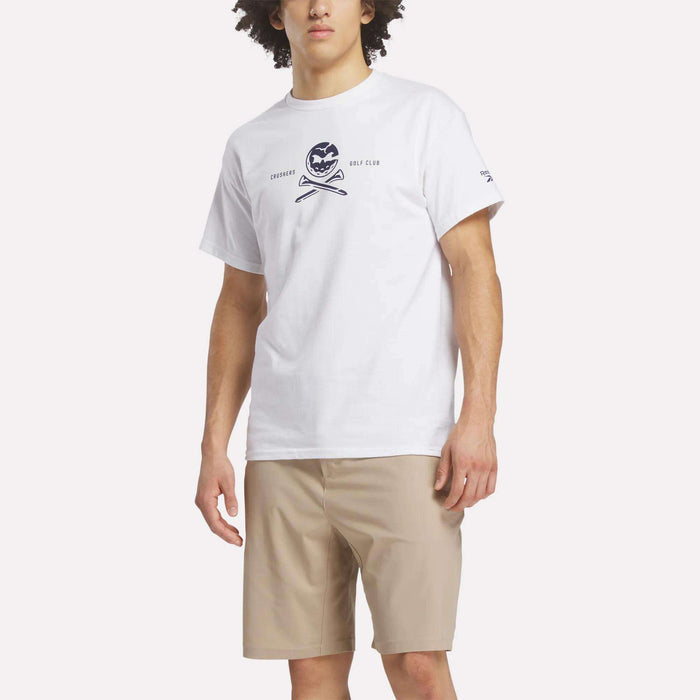A man wears the Reebok Men's Crushers Tee in white, featuring a logo and text, with beige shorts against a plain light background.