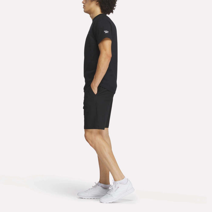A man stands in profile with hands in pockets, wearing the Reebok Men's Crushers Tee in black, paired with black shorts and white sneakers against a plain white background.