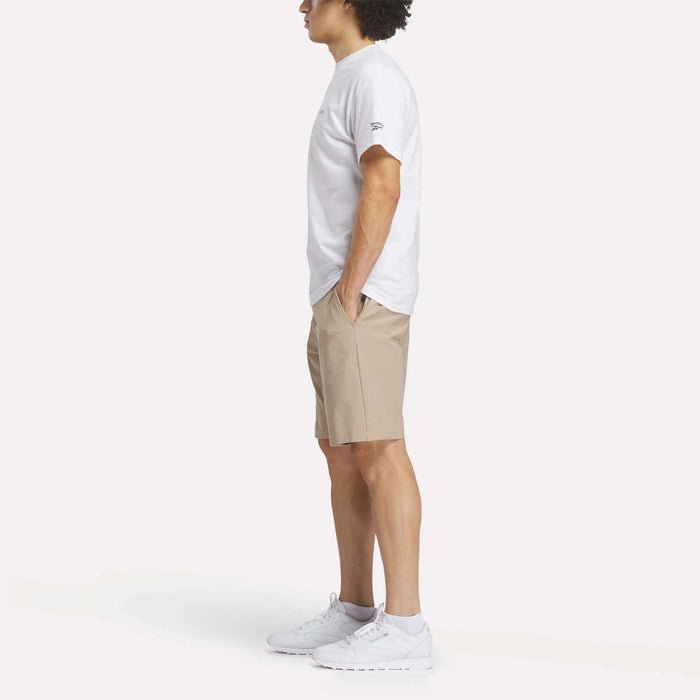 A person stands in profile with hands in pockets, wearing a white Reebok Men's Crushers Tee, tan shorts, and white sneakers against a plain white background.