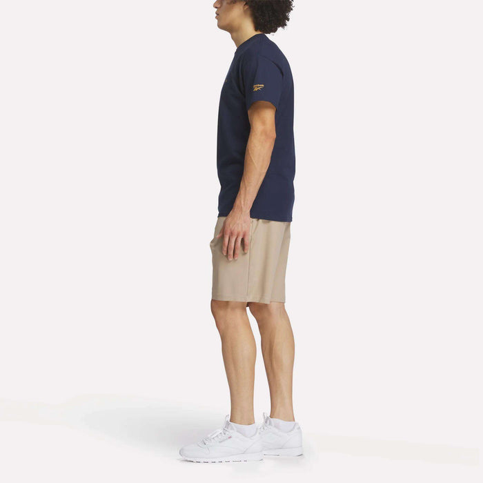 A man stands in profile against a plain white background wearing the Reebok Men's Crushers Tee, beige shorts, and white sneakers.