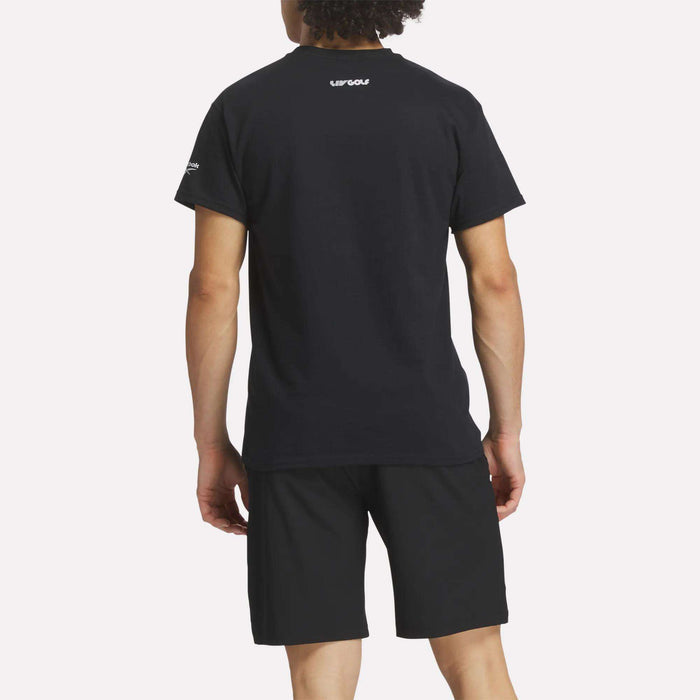 A person stands with their back to the camera, wearing a black Reebok Men's Crushers Tee and black shorts. The tee features small white text on the upper back and left sleeve, set against a plain light background.