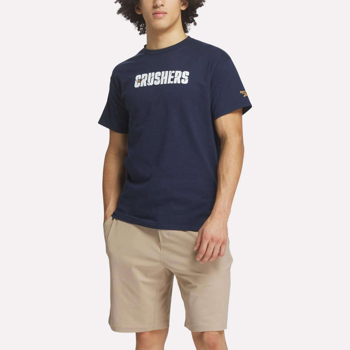 A man wearing a Reebok Men's Crushers Wordmark Tee in navy blue and beige shorts stands against a plain white background.