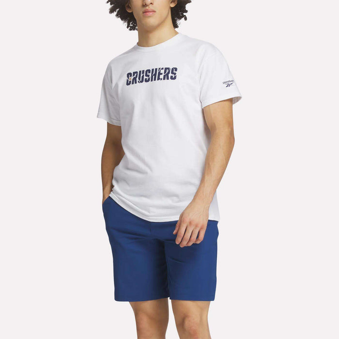 A person wears the Reebok Men's Crushers Wordmark Tee and blue shorts, standing with one hand in their pocket against a plain background, emulating Bryson DeChambeau's sporty style.