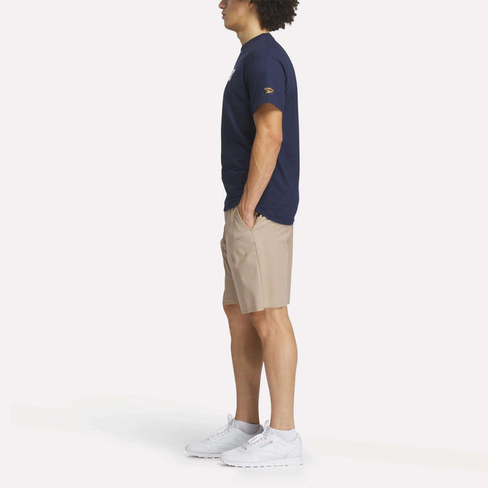A person stands in profile with hands in pockets, wearing a Reebok Men's Crushers Wordmark Tee in navy, beige shorts, and white sneakers against a plain white background.
