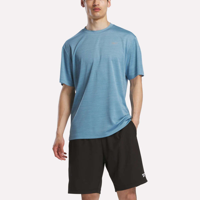A man wears a Reebok Men's Director Short Sleeve Top and black shorts, standing with one hand in his pocket against a plain white background.