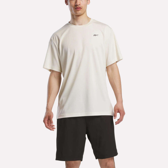 A person wears a loose-fitting Reebok Men's Director Short Sleeve Top with black shorts, standing against a plain light background.