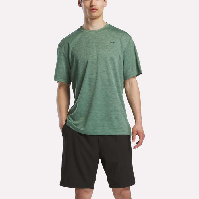 A person in a green Reebok Men's Director Short Sleeve Top and black shorts stands against a plain white background.