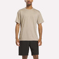 A man stands facing forward wearing a Reebok Men's Director Short Sleeve Top in Grainy Grey Heather and black shorts against a plain white background.