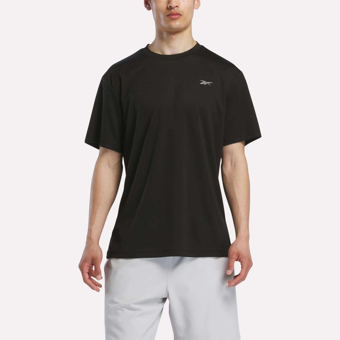 A man stands against a plain white background wearing Reebok's Men's Director Short Sleeve Top in black and white shorts. The Reebok logo appears on the upper left of this stretchy training shirt.