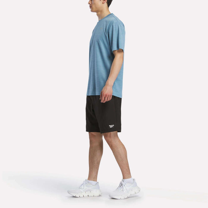 A person in a light blue Reebok Men's Director Short Sleeve Top, black shorts, and white athletic shoes stands in profile against a plain white background.