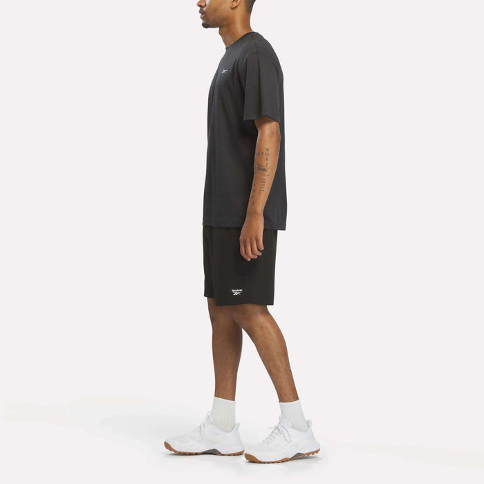 A man stands in profile wearing a black Reebok Men’s Director Short Sleeve Top, black shorts, white socks, and white sneakers against a plain white background.