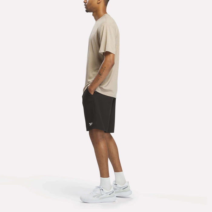 A person stands in profile wearing the Reebok Men's Director Short Sleeve Top in beige, along with black shorts, white socks, and white sneakers against a plain white background.