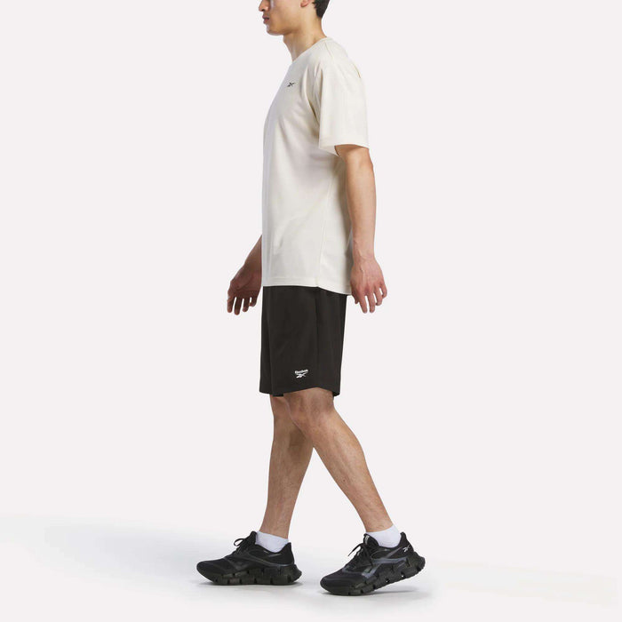 A man wearing a Reebok Men's Director Short Sleeve Top, black shorts, and black sneakers walks left against a plain white background.