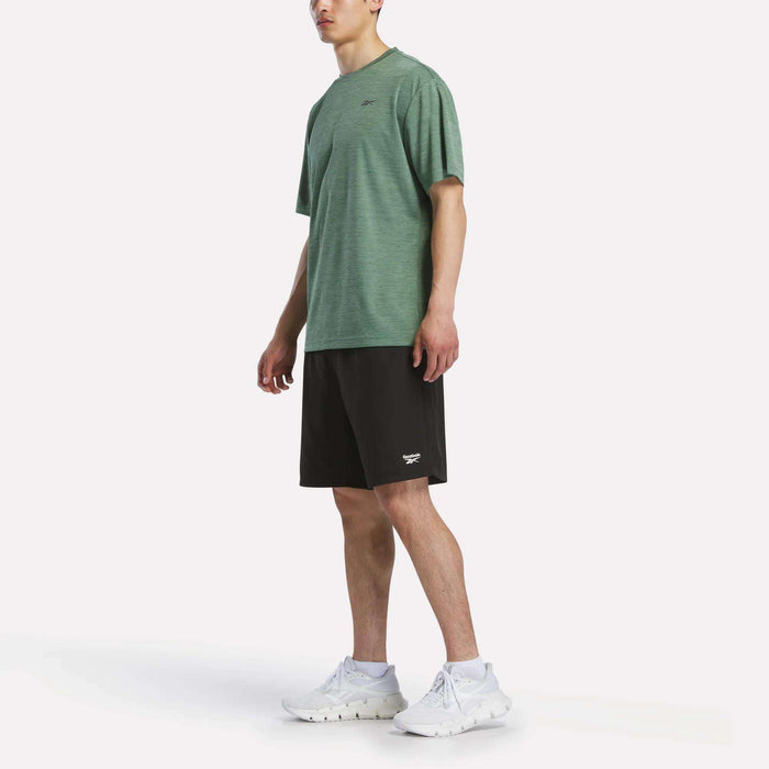 A person stands against a plain white background wearing a green Reebok Men's Director Short Sleeve Top, black shorts, and white athletic shoes. The look is sporty and casual. The person's face is partially out of frame.