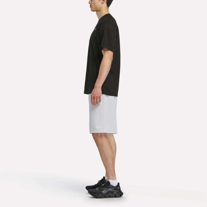 A man stands in profile facing left, wearing a Reebok Men's Director Short Sleeve Top, light gray athletic shorts, black sneakers, and white socks against a plain white background.