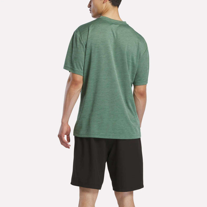 A man stands facing away, wearing a green Reebok Men's Director Short Sleeve Top and black shorts against a plain white background.