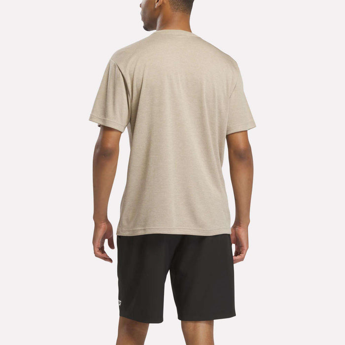 A man stands facing away, wearing the Reebok Men's Director Short Sleeve Top in beige with stretchy short sleeves and black shorts against a plain white background.