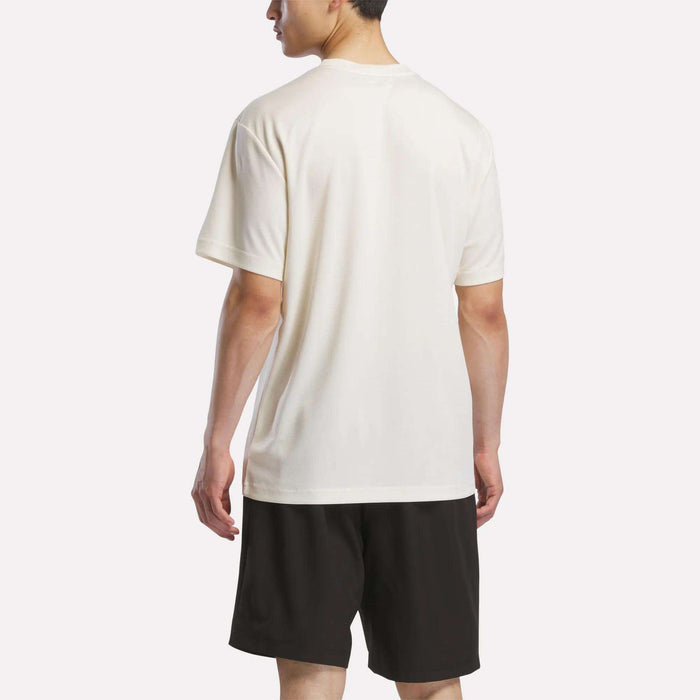 A man faces away from the camera wearing the Reebok Men's Director Short Sleeve Top in white and black shorts, set against a plain light background.