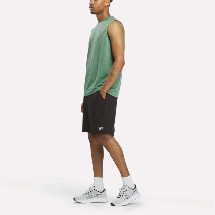 A person stands sideways against a plain white background, wearing the Reebok Men's Elite Sleeveless Top in green—crafted from cotton flex knit—paired with black shorts, white crew socks, and white Reebok athletic shoes.