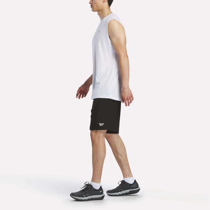 A man in a Reebok Men's Elite Sleeveless Top, black athletic shorts, white socks, and black sports shoes walks left against a plain white background. The cotton flex knit fabric delivers comfort and style.