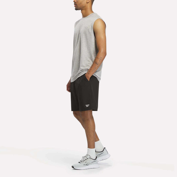 A man wearing a Reebok Men's Elite Sleeveless Top in gray, black shorts, white socks, and white sneakers stands with one hand in his pocket against a plain white background.