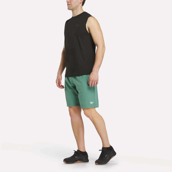 A man in a Reebok Men's Elite Sleeveless Top, green athletic shorts, and black sneakers stands turned slightly to the side against a plain white background.