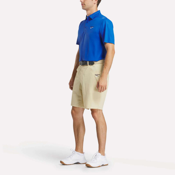 A man stands facing slightly to the side, wearing Reebok Men's Favorite 5-Pocket Golf Shorts 9" along with a blue polo, black belt, and white athletic shoes against a plain white background.
