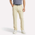 A man stands with his hands in his pockets, wearing light beige Reebok Men's Favorite 5-Pocket Relaxed Fit Golf Pants featuring RBK-DRY tech, a black belt, gray polo, and white sneakers. The image is cropped at the shoulders; his face is not visible.