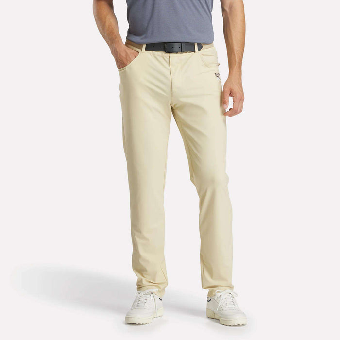 A man stands with his hands in his pockets, wearing light beige Reebok Men's Favorite 5-Pocket Relaxed Fit Golf Pants featuring RBK-DRY tech, a black belt, gray polo, and white sneakers. The image is cropped at the shoulders; his face is not visible.