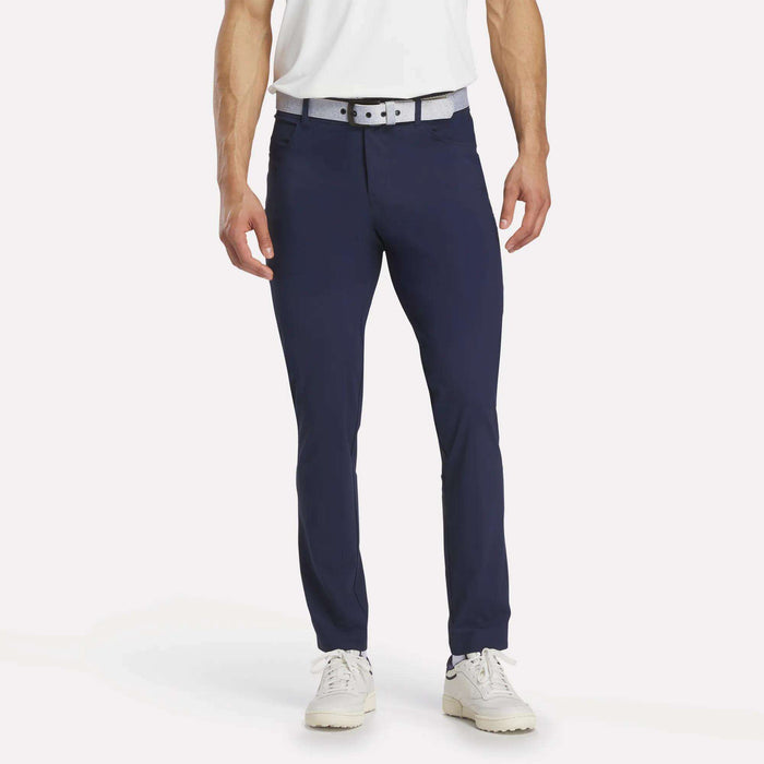 A person wearing a white shirt, white and gray belt, and white shoes stands against a plain white background, modeling Reebok Men's Favorite 5-Pocket Relaxed Fit Golf Pants in navy blue.