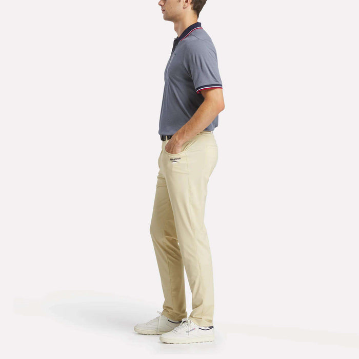 A man stands in profile wearing a short-sleeved blue polo shirt, men's Reebok Favorite 5-Pocket Relaxed Fit Golf Pants, and white sneakers against a plain white background.