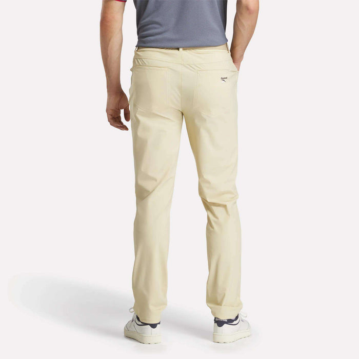 A person is seen from the back wearing Reebok Men's Favorite 5-Pocket Relaxed Fit Golf Pants in light beige with RBK-DRY tech, a gray short-sleeve shirt, and white sneakers, posed against a plain white background.