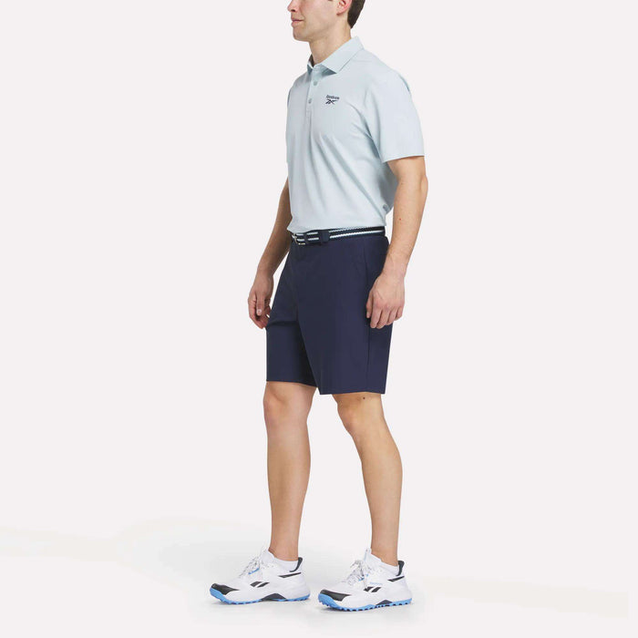 A man stands against a white background wearing a light blue polo, a black belt, white athletic shoes with blue accents, and Reebok Men's Favorite Golf Shorts 8.5" in navy.