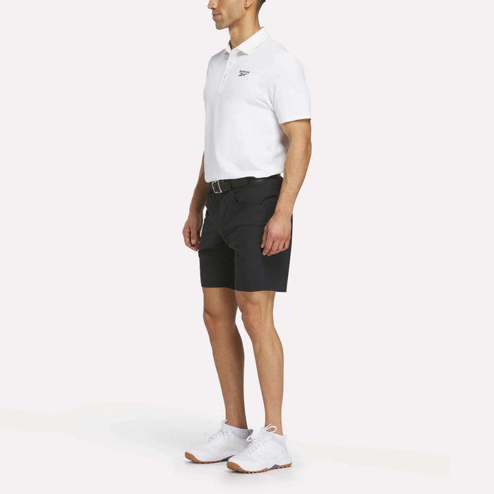 A man stands slightly turned to the side, wearing a white polo, Reebok Men's Favorite Golf Shorts 8.5" in black with a belt, and white sneakers against a plain white background.