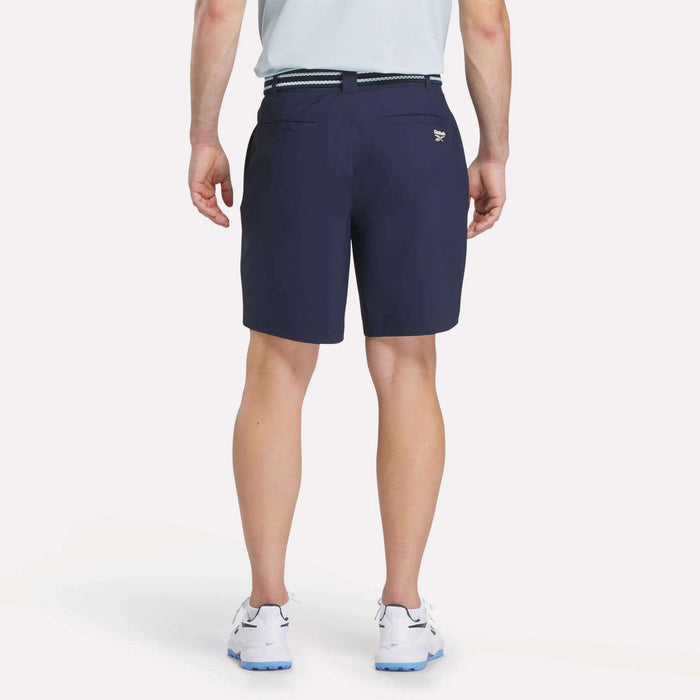 A man faces away, wearing a light blue shirt and Reebok Men's Favorite Golf Shorts 8.5" in navy with a belt, paired with white athletic shoes. The background is plain and light-colored.