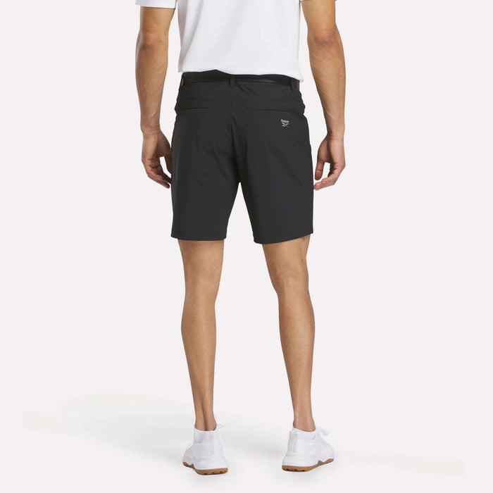 A person stands facing away from the camera, wearing a white polo shirt, Reebok Men's Favorite Golf Shorts 8.5", and white sneakers against a plain background.