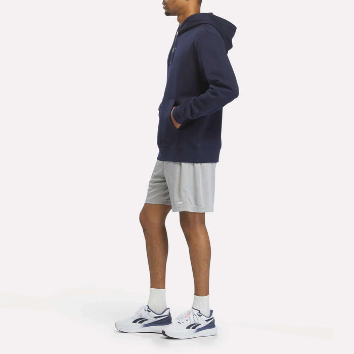 A person stands in profile wearing a navy Reebok Men's Finisher Hoodie, gray shorts, white socks, and white sneakers with blue accents against a plain light background.