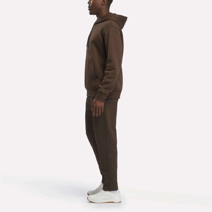 A person stands in profile wearing the Reebok Men’s Finisher Hoodie paired with matching joggers and white sneakers against a plain white background.