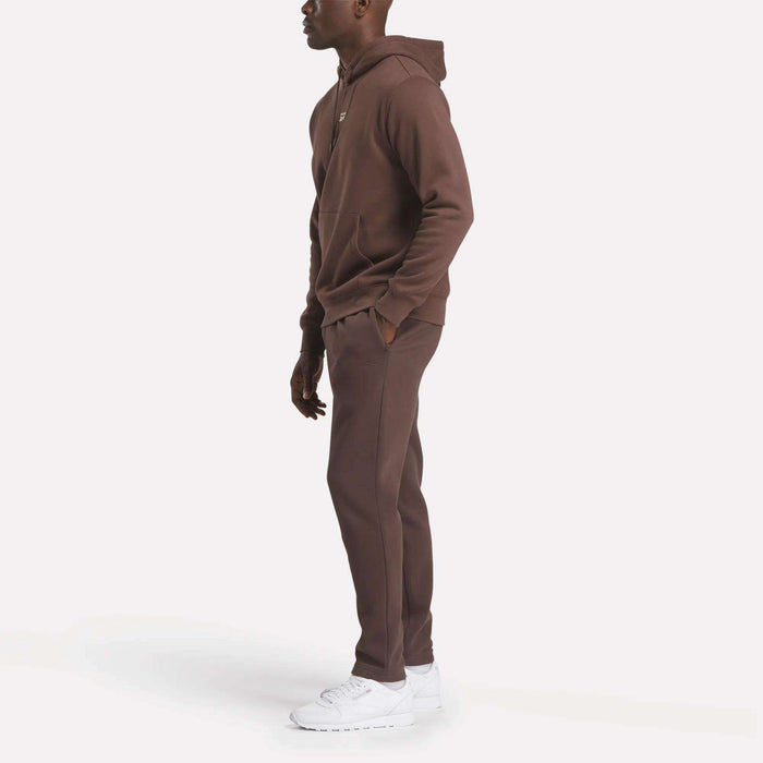 A person stands sideways in a Reebok Men's Finisher Hoodie and matching sweatpants with white sneakers, set against a plain white background.