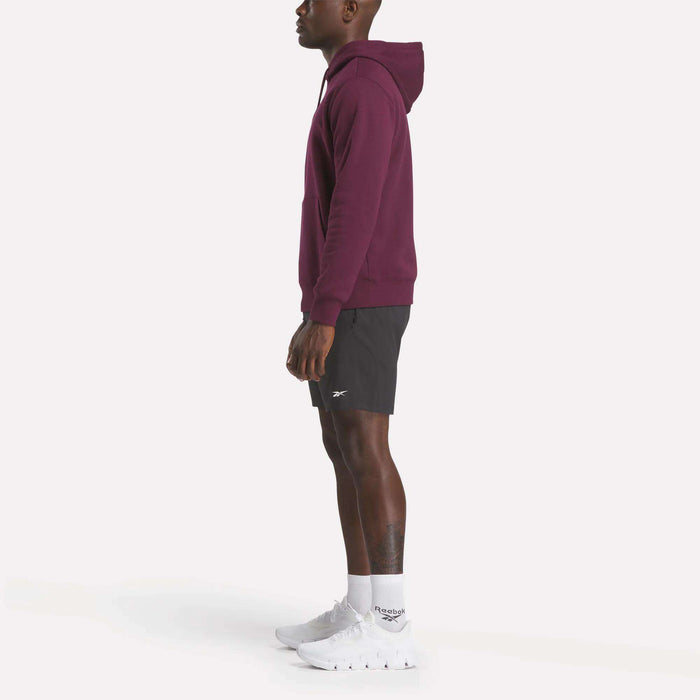 A man stands in profile wearing the Reebok Men's Finisher Hoodie in burgundy, paired with black athletic shorts, white socks, and white sneakers against a plain white background.