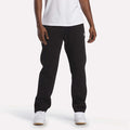 A person stands against a plain white background, with only the lower half of their body visible, showcasing the cozy comfort of the Reebok Men's Fleece Straight Leg Pants in black. They are styled here with a white t-shirt and white sneakers.