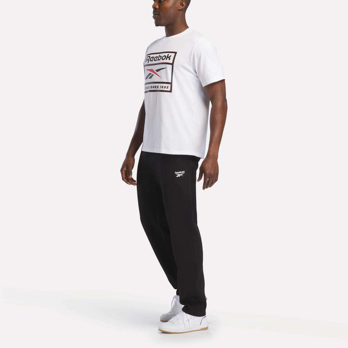 A man stands against a plain white background wearing a white Reebok t-shirt, Men's Fleece Straight Leg Pants from Reebok in black for cozy comfort, and white sneakers.