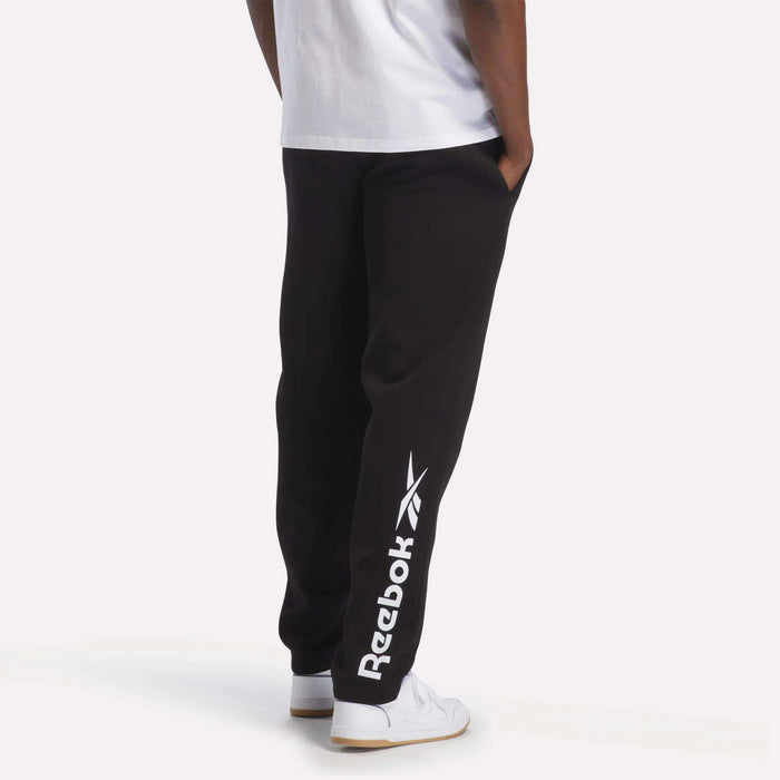 A man is wearing Reebok Men's Fleece Straight Leg Pants in black, featuring a white logo, paired with a white T-shirt and white sneakers. He stands with his hands in his pockets against a plain white background. These pants are crafted from cozy brushback fleece for all-day comfort and classic athletic style.