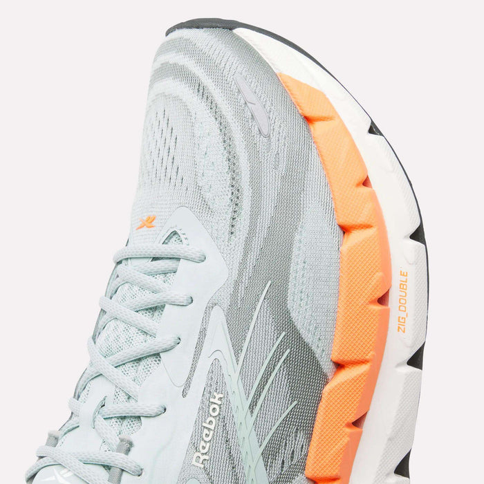 Close-up of the Reebok Men's FloatZig Double Running Shoes, featuring a light gray breathable mesh upper, laces, and orange accents on the thick, jagged sole with "Zig Double" text.