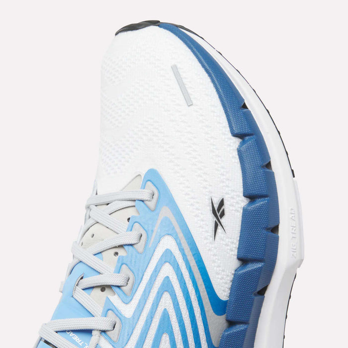 Close-up of Reebok Men's FloatZig Tread Running Shoes in white and blue, featuring a wavy upper design, gray laces, black logo, and a thick segmented dual-density midsole—ideal for treadmill workouts or daily runs.
