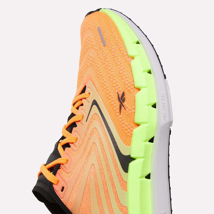 A close-up of Reebok Men's FloatZig Tread Running Shoes with an orange knit upper, black accents, and a bright green segmented sole. The cushioned midsole and wavy design offer comfort and style—ideal for treadmill workouts.